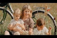 Mutter mit zwei Töchtern sitzt im Feld vor abgestelltem Fahrrad und pflückt Blumen