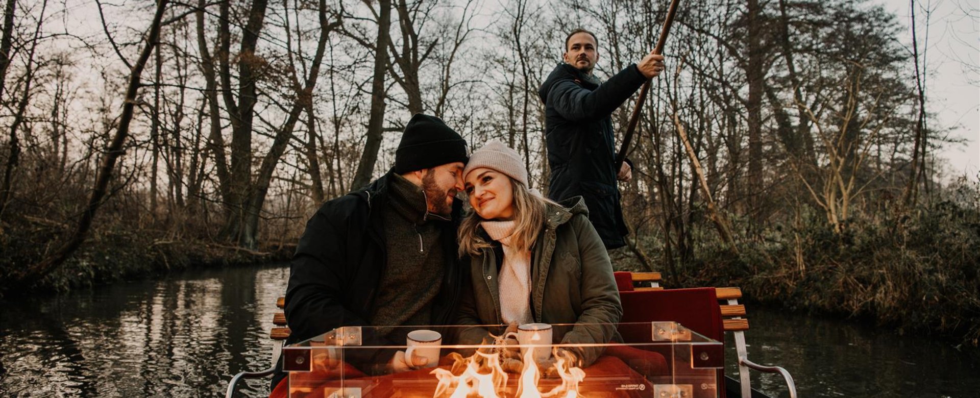 Ein Paar sitzt im Winter auf einem Kahn vor einem Kaminfeuer und wird durch den Spreewald gefahren.