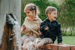 Two children girl and boy sit on the edge of a fountain and laugh while water splashes up