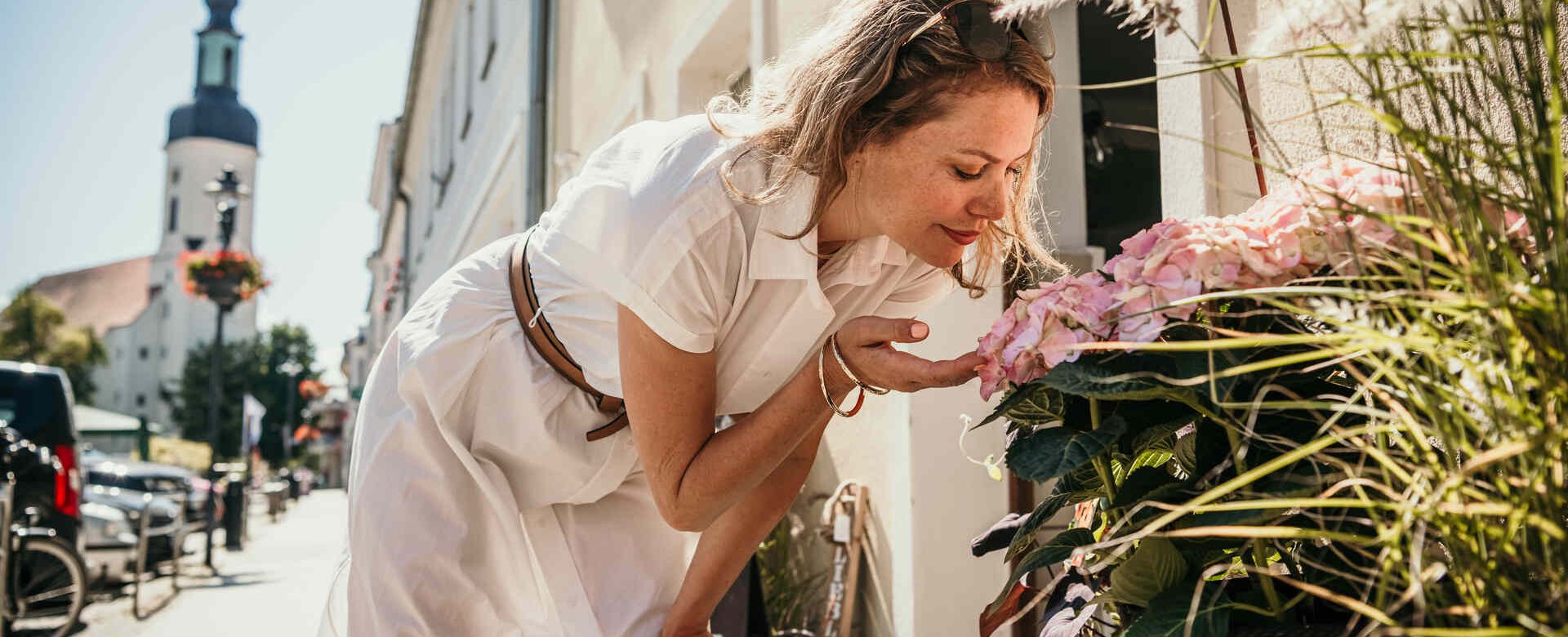 Frau steht vor Blumenladen und riecht an Blume im Hintergrund eine Stadtszene mit Kirche
